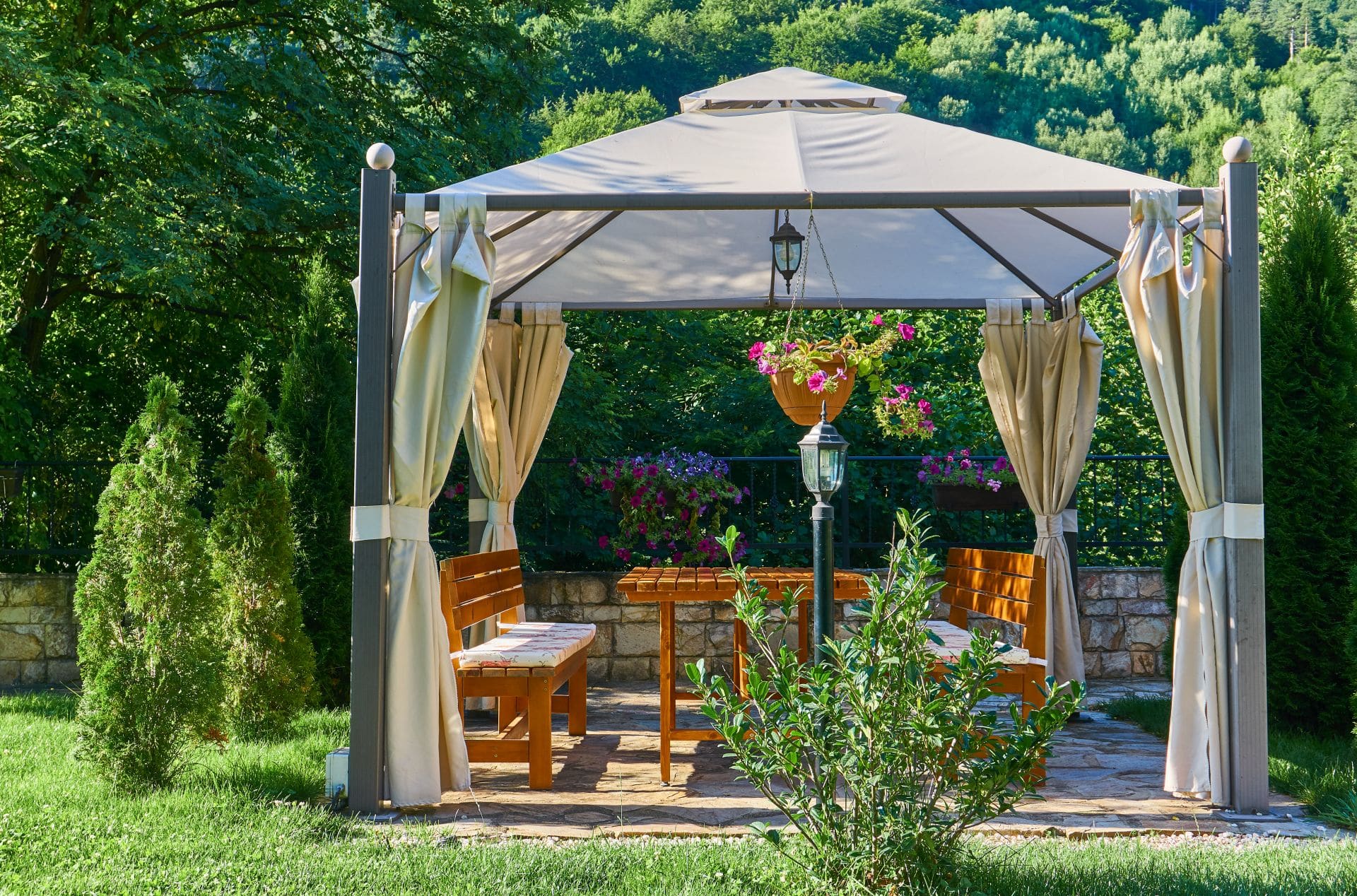 Ein eleganter Pavillon im Garten mit Vorhängen, einer Holz-Sitzgruppe und dekorativen Pflanzen, ideal für entspannte Stunden im Freien.